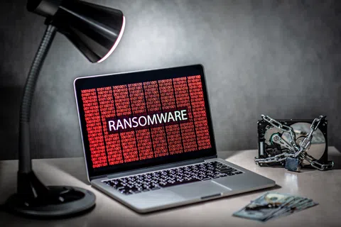 Laptop displaying ransomware warning in red binary code with chained hard drives and desk lamp in dim room.