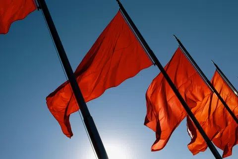 Bright red flags waving on poles against a clear blue sky with sunlight behind them